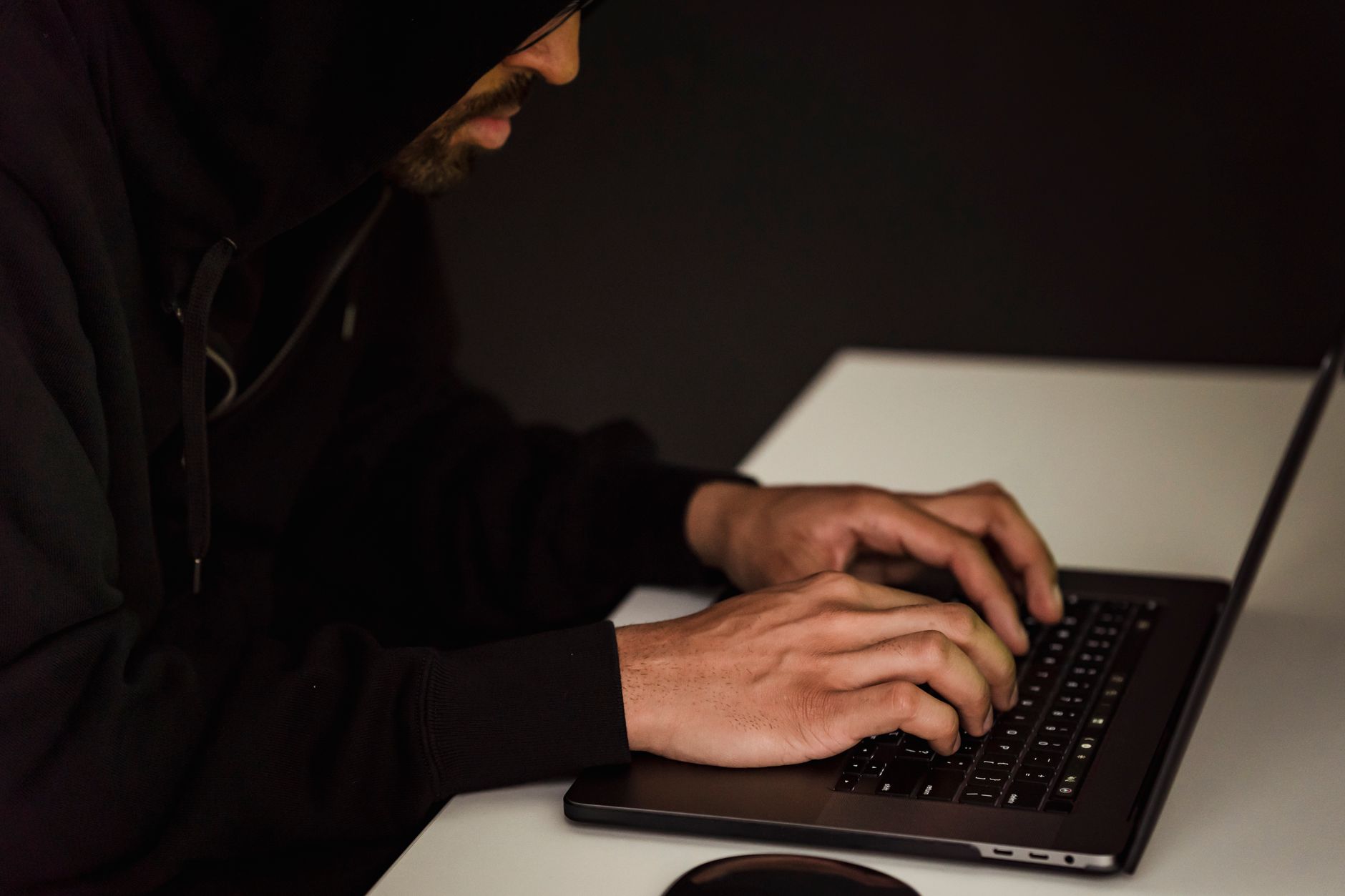 Mężczyzna w czarnej bluzie z kapturem przy laptopie w ciemnym pokoju